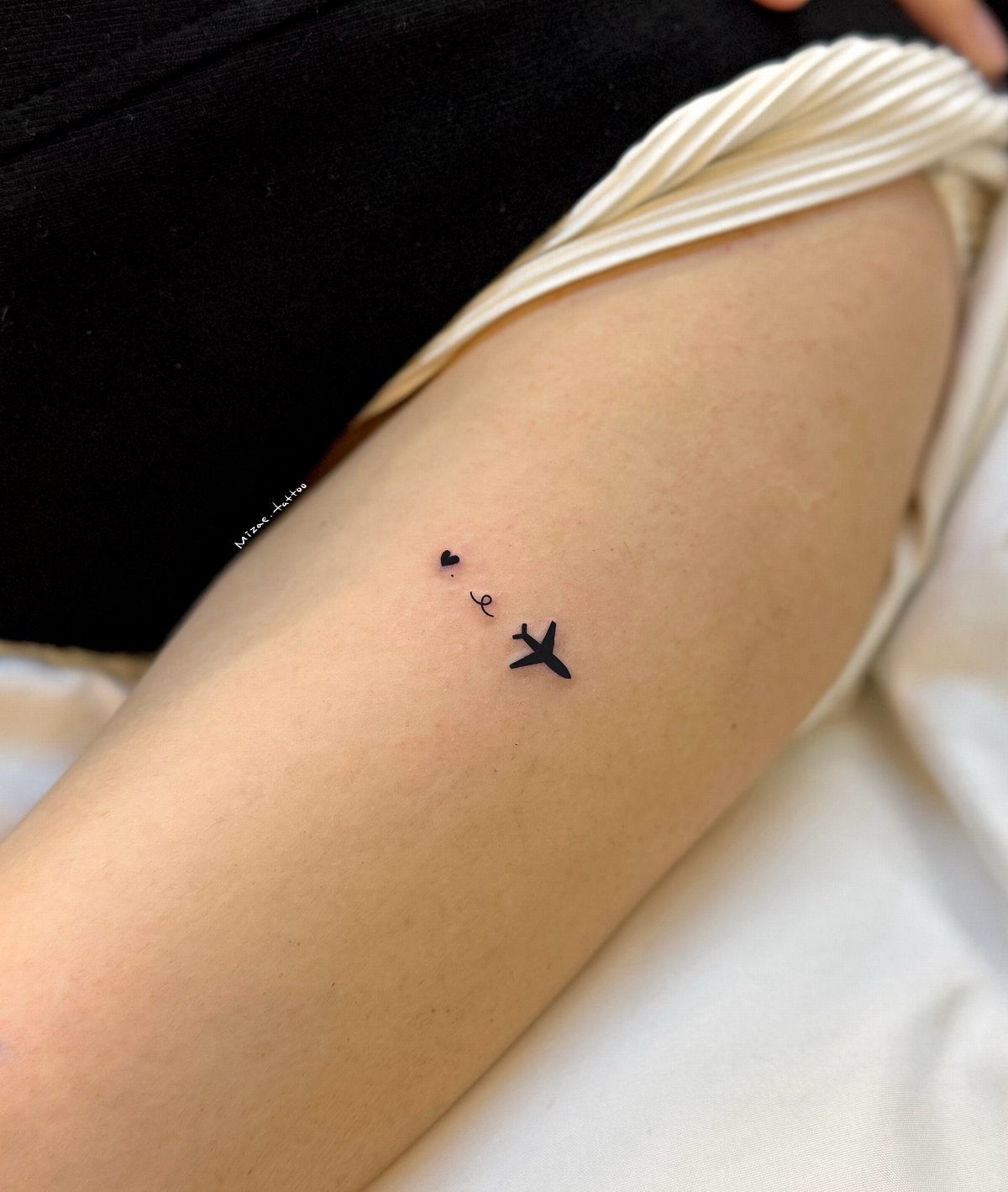 Small black paper airplane tattoo with a dotted trail and heart on skin.