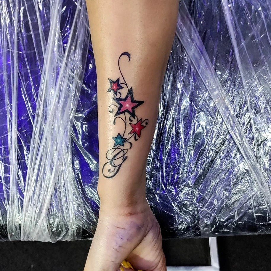Colorful star tattoos with a swirling line and letter G on a forearm.