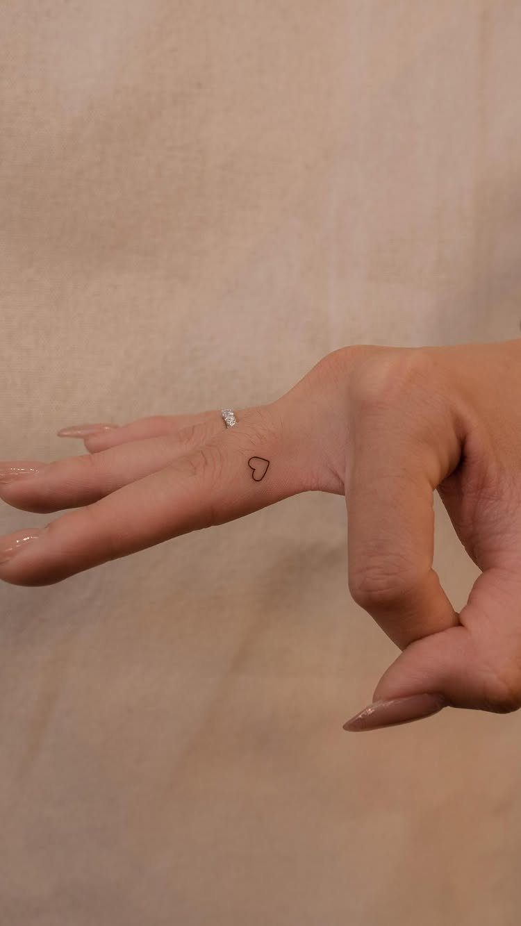 Small black heart tattoo on the side of a woman's ring finger.