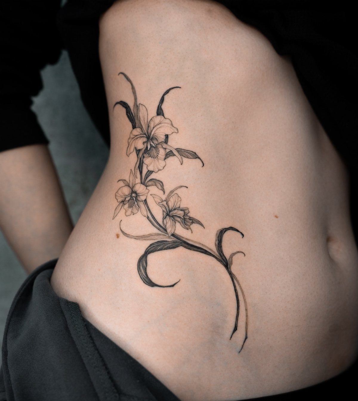 Cascading orchid flower tattoo in black ink on a woman's ribcage and side.