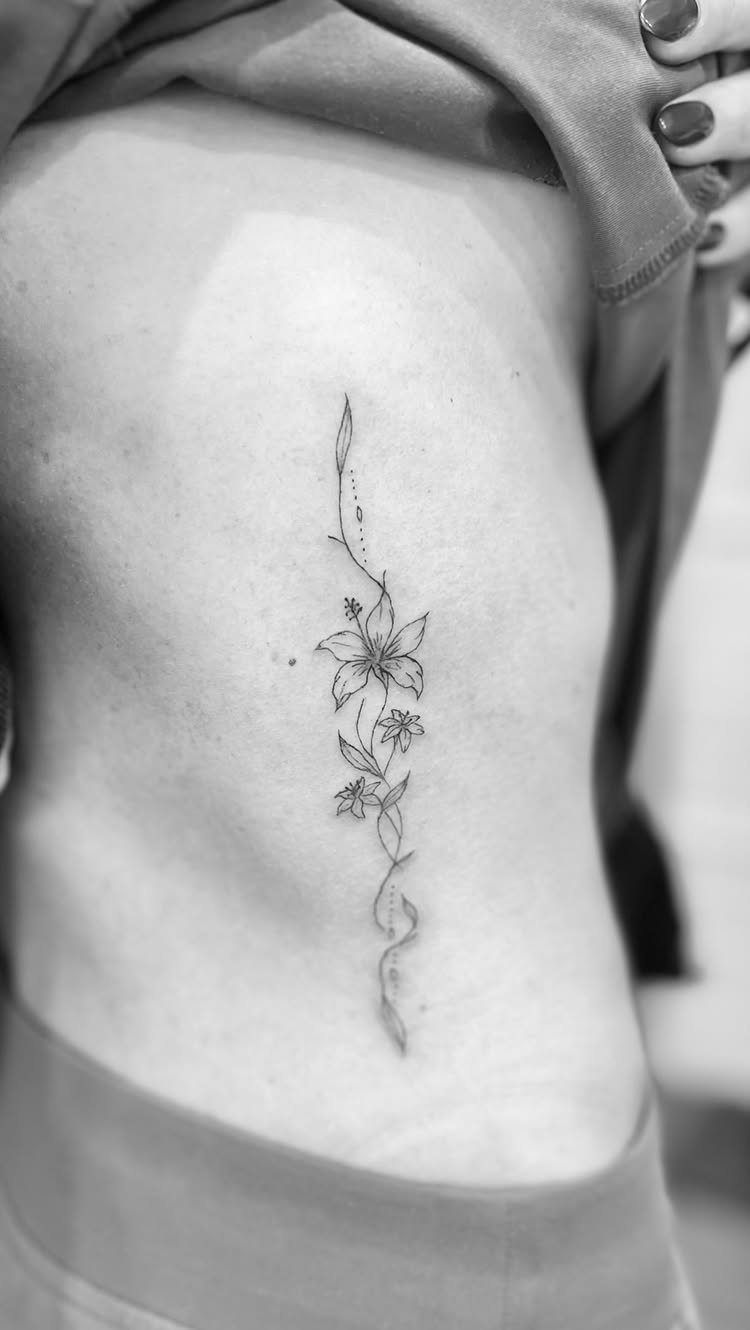 Lily and wildflower tattoo cascading vertically down a woman's ribcage in black ink.