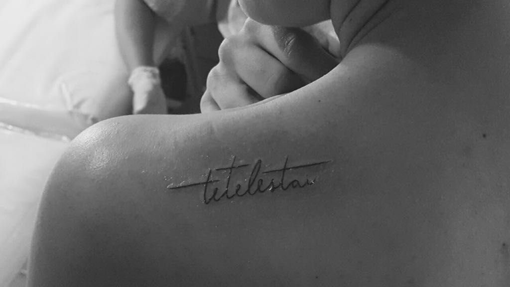 Cursive script tattoo reading tetelestai on a person's upper arm and shoulder.