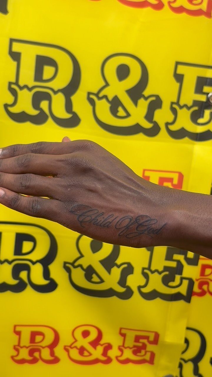 Hand with a cursive Child Of God tattoo against a yellow patterned background