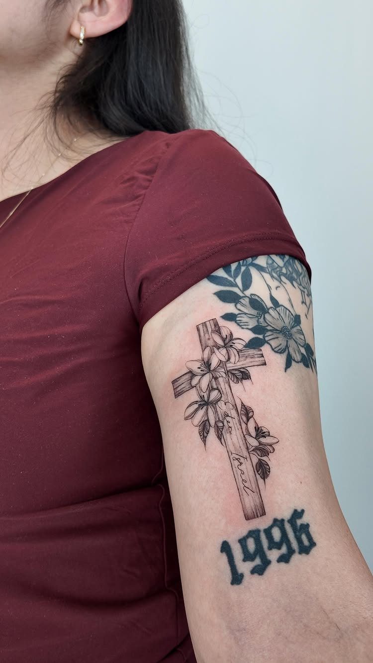 Floral cross tattoo on an upper arm with the year 1996 below it.