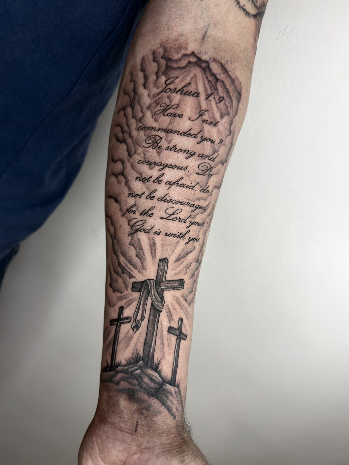 Forearm tattoo featuring Joshua 1:9 scripture above three crosses on a hill.