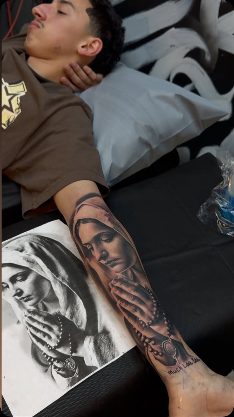 Black and grey tattoo of Virgin Mary on a forearm next to reference drawing