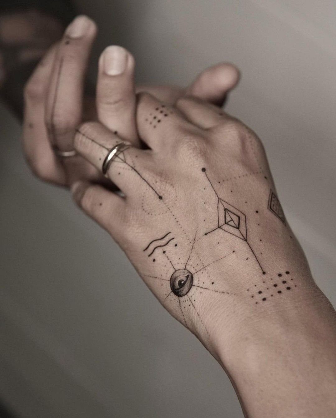 Hand and fingers with fine-line geometric tattoos, dots, and a small eye design.