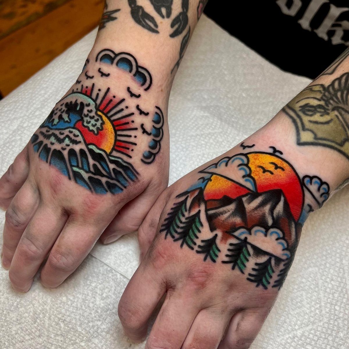Two hands with colorful traditional tattoos of mountains, waves, and a setting sun.