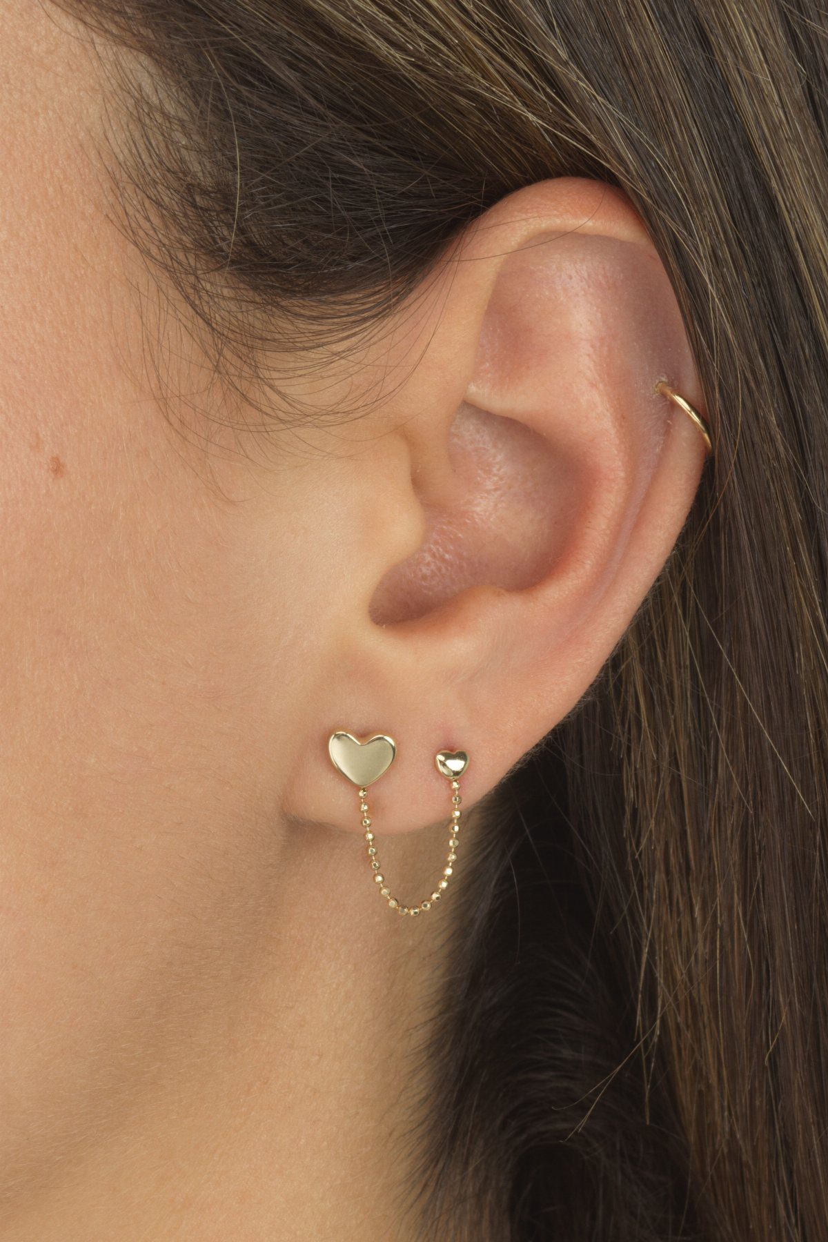 Hanging Ear Piercing Ideas: 6 Chain, Pearl and Star Styles 2 Gold heart stud earrings connected by a draped chain on a person's ear.