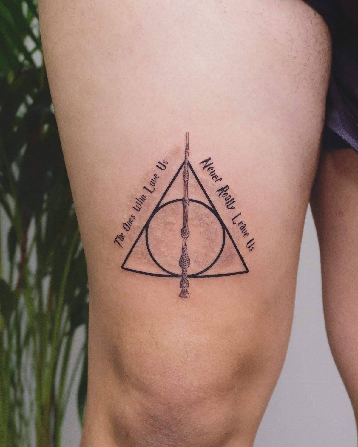 Deathly Hallows tattoo on a thigh with a sentimental quote in cursive text.