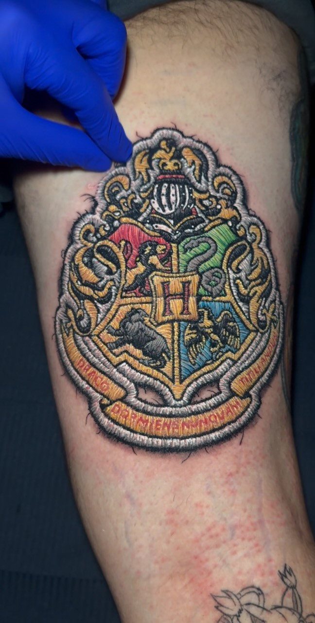 Hogwarts crest tattoo on skin with colorful house symbols and a central H