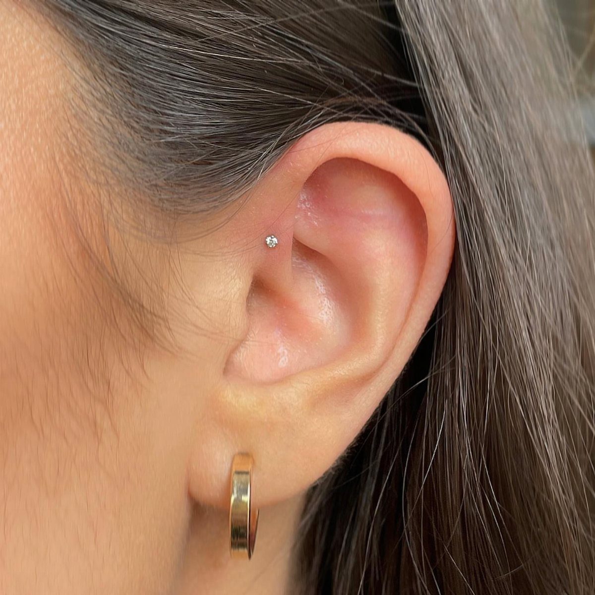 Ear with a silver stud in the forward helix and a gold hoop earring.