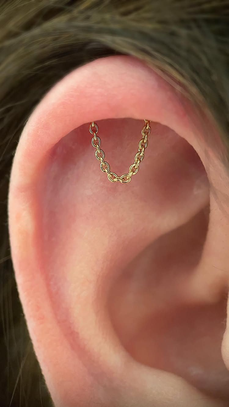Human ear with two gold helix piercings connected by a delicate gold chain.