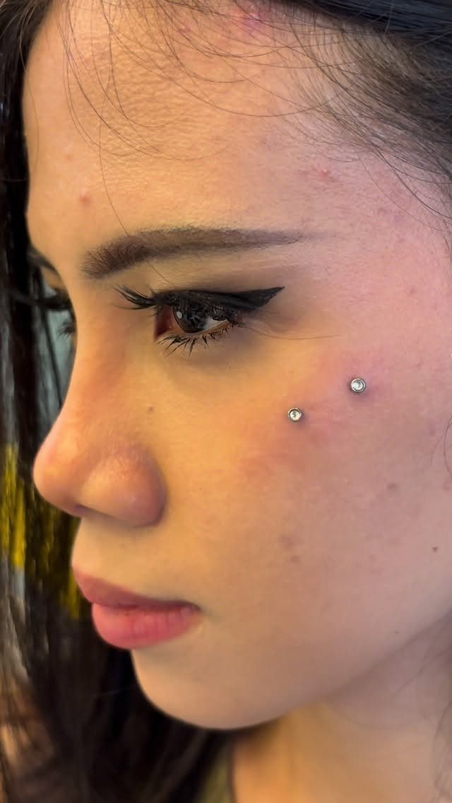 Person's face with two silver stud anti-eyebrow piercings on the upper cheekbone.