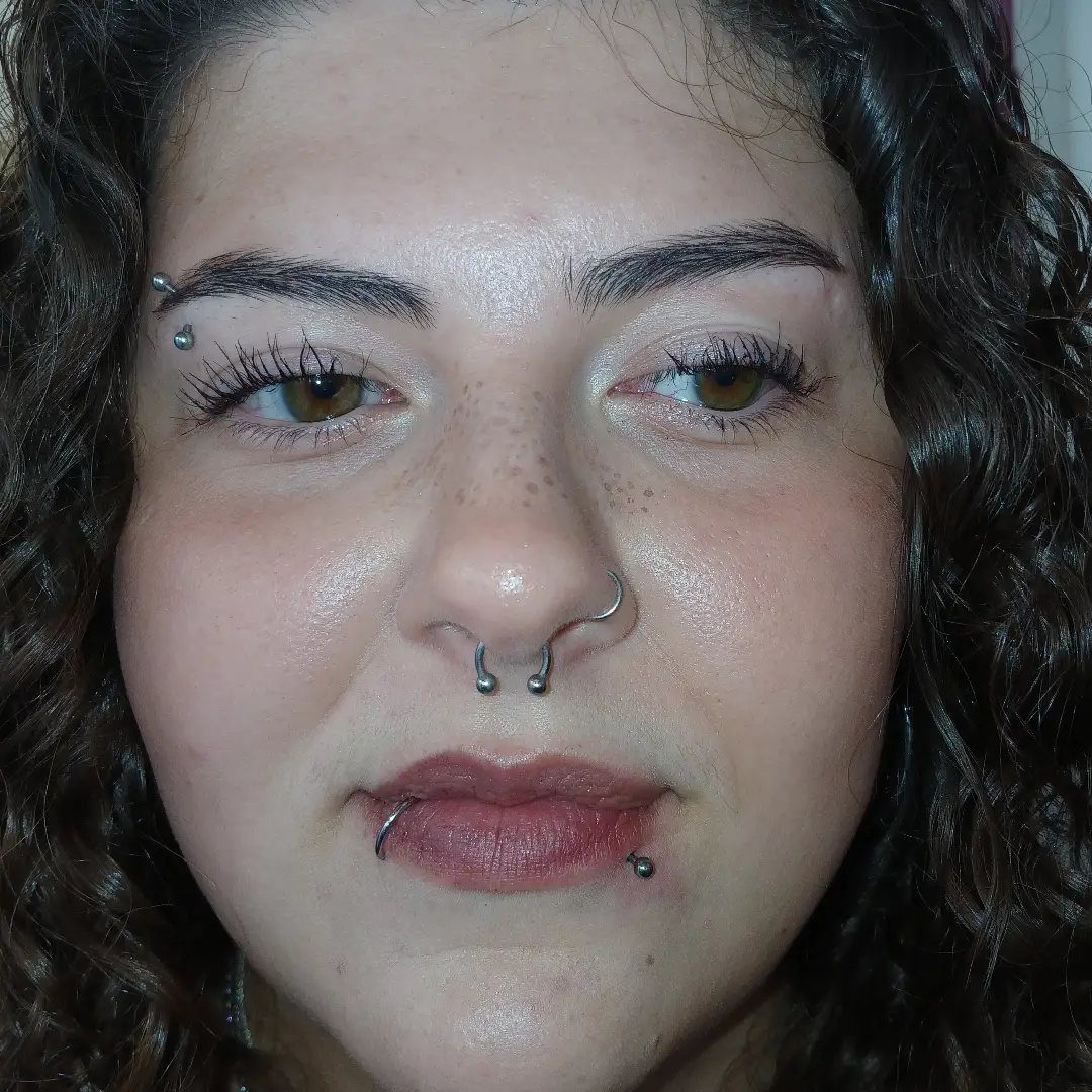 Person with curly hair featuring eyebrow, septum, and lip piercings.