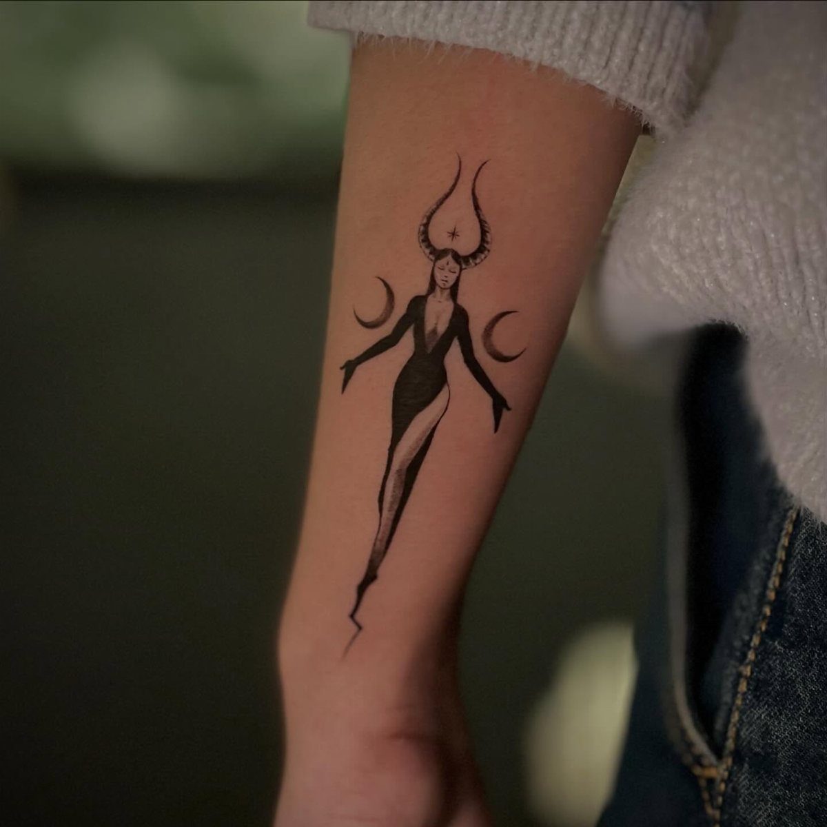 Black ink tattoo of a horned female figure with crescent moons on a forearm