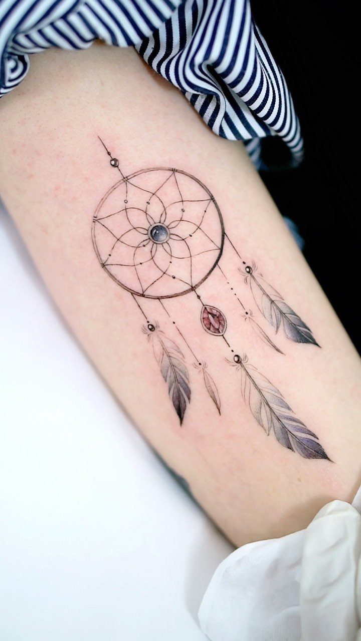 Dreamcatcher tattoo on a forearm with detailed webbing and hanging grey feathers.