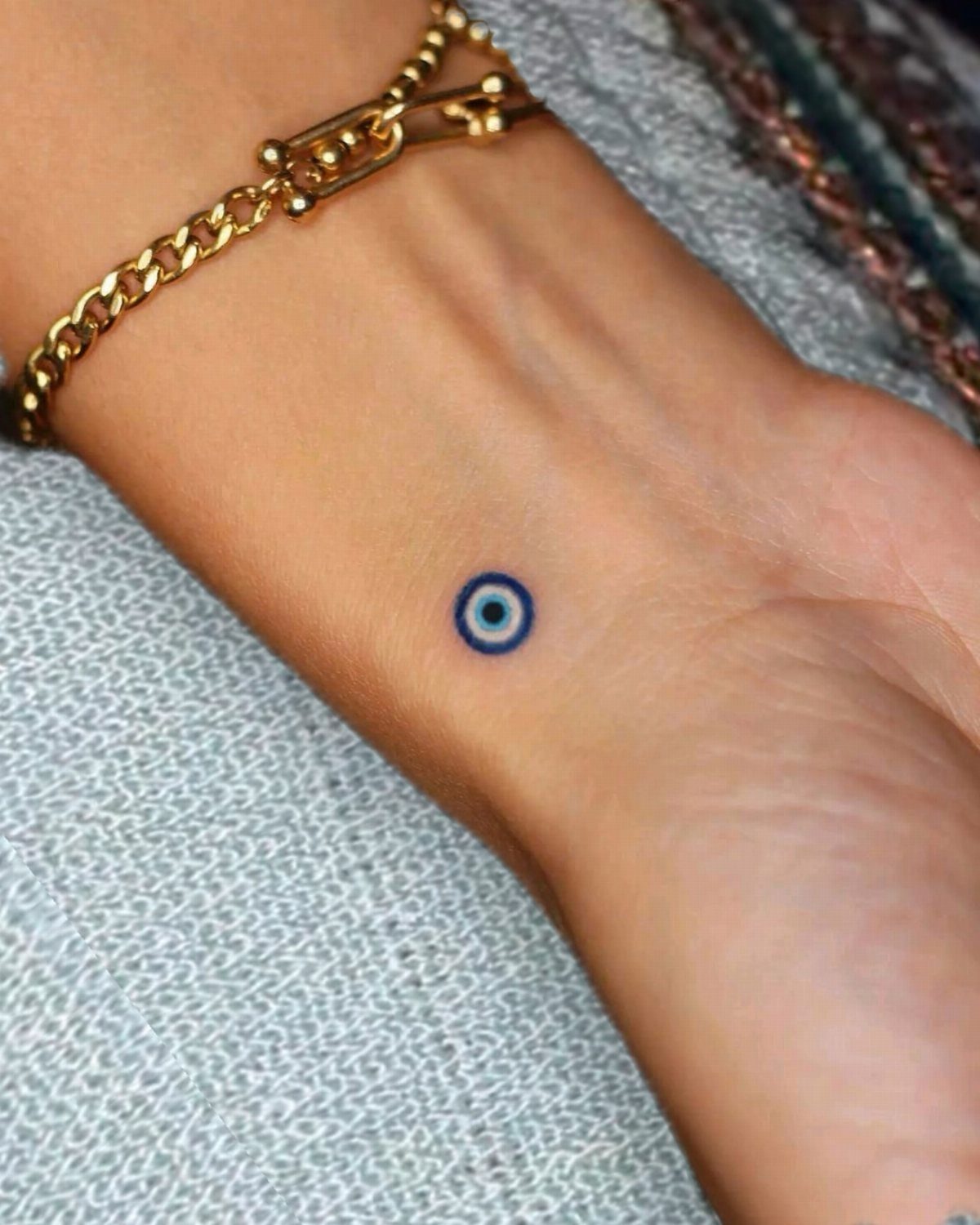 Small blue and white evil eye tattoo on a person's wrist with gold bracelet.