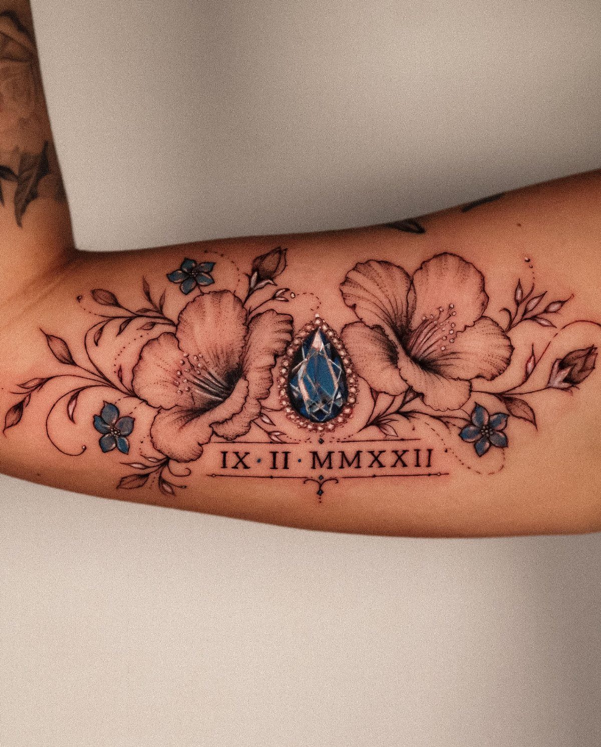 Tattoo on a forearm featuring a blue gemstone surrounded by white flowers and Roman numerals.