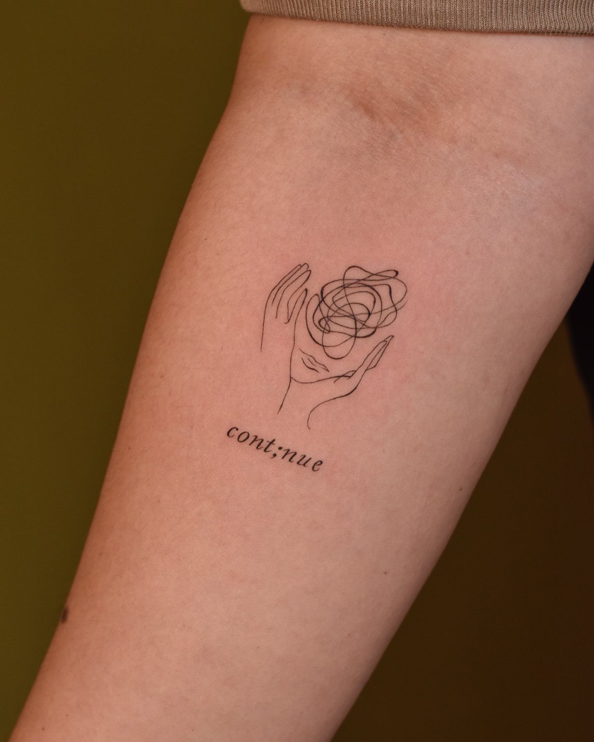 Tattoo of a hand holding a scribble with the word cont;nue on an arm.