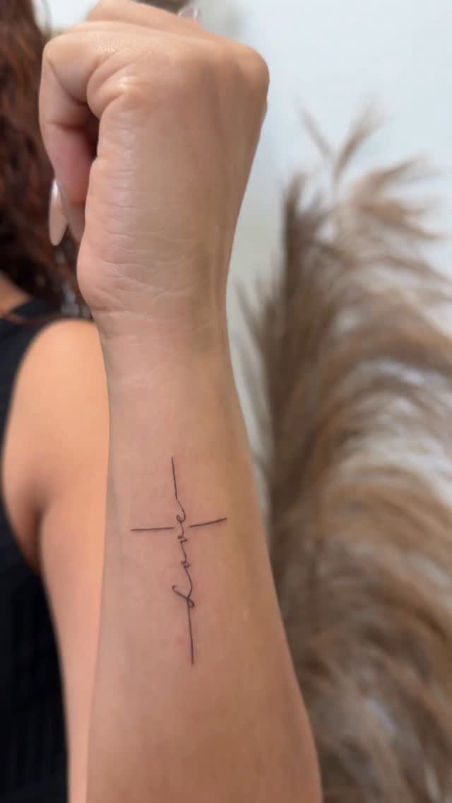 Minimalist Fine Line Tattoo Ideas: 9 Styles to Consider 3 Small fine line cross tattoo with the word love on a person's wrist.