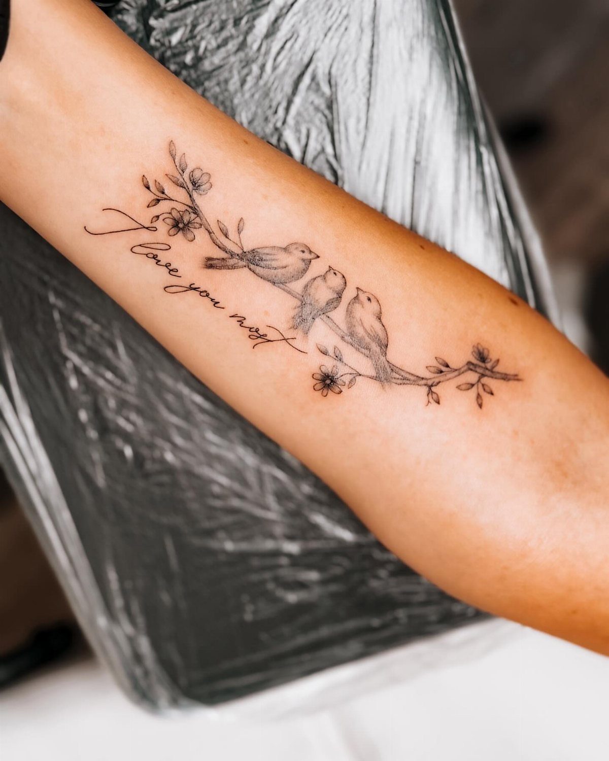 Three birds on a flowering branch tattoo with cursive text on a forearm