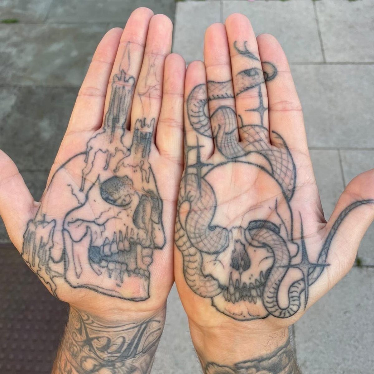 Two open palms with large black ink skull tattoos on a stone background.