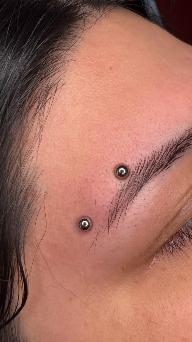 Piercing Between Eyebrows: 8 Styles and Placements 1 Horizontal eyebrow surface piercing with two silver beads on a person's face.