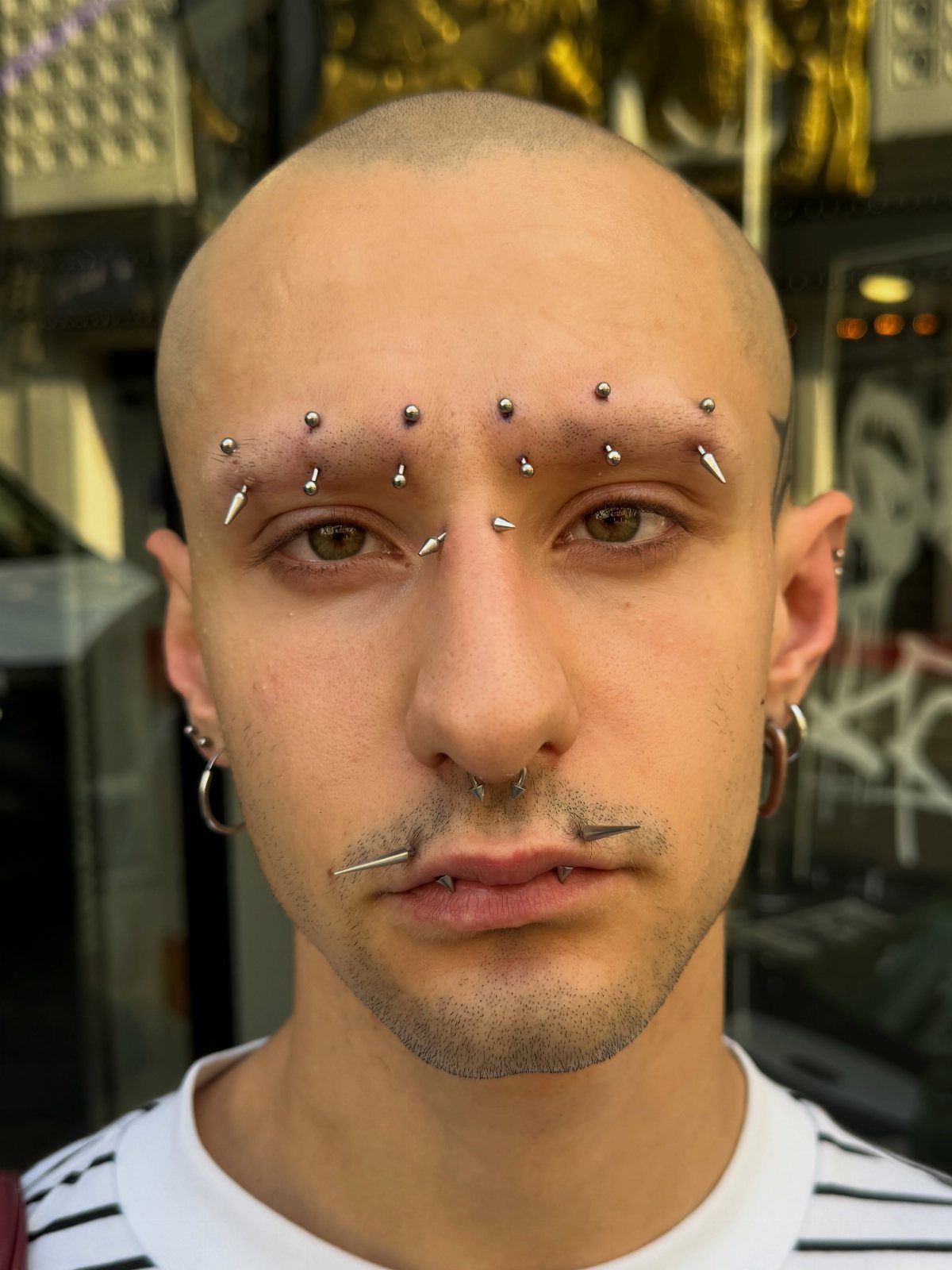 Piercing Between Eyebrows: 8 Styles and Placements 4 Bald man with multiple silver piercings across his eyebrows, nose, and lips.