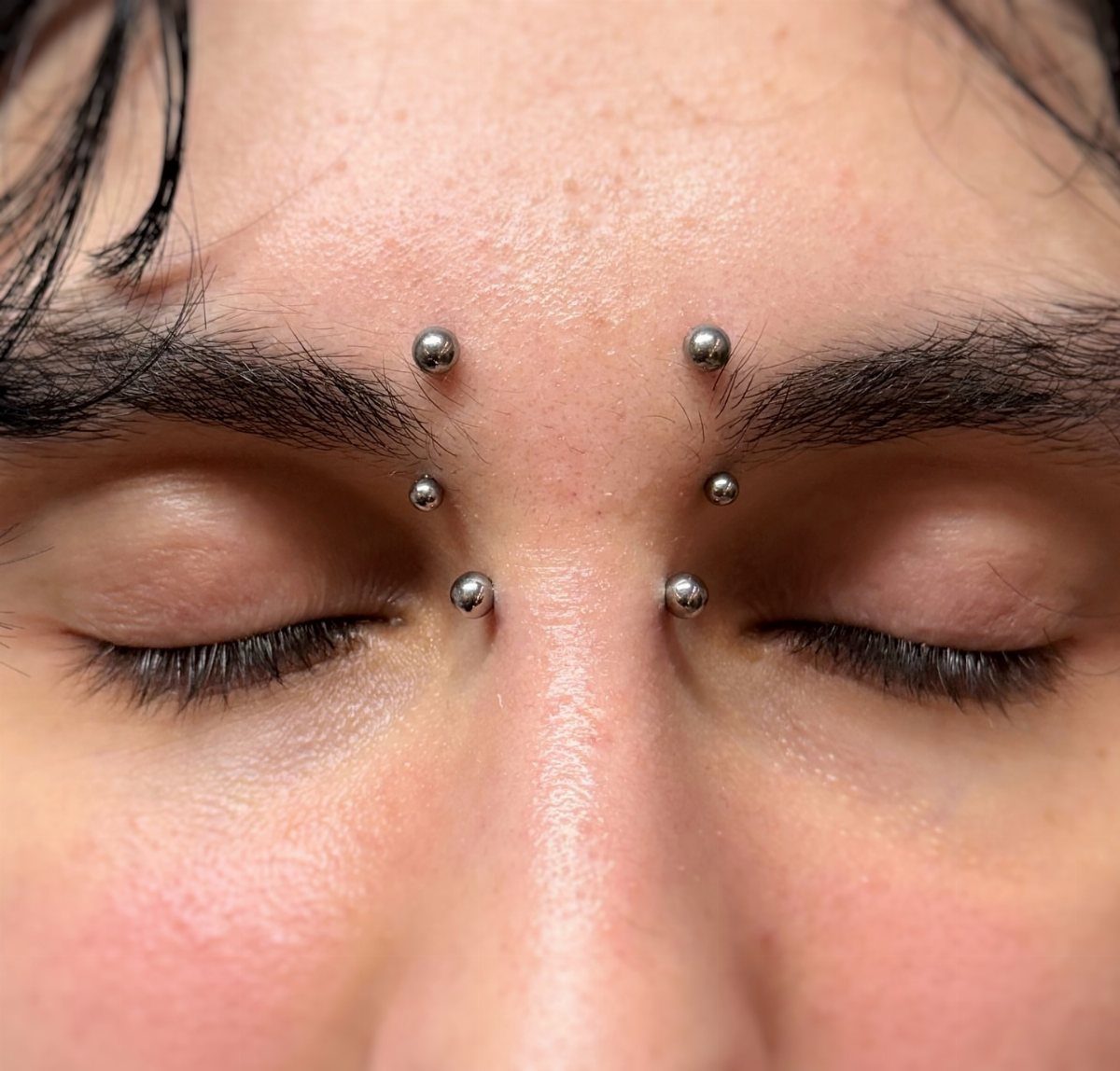 Piercing Between Eyebrows: 8 Styles and Placements 7 Six silver ball piercings arranged in two columns between a person's eyebrows.