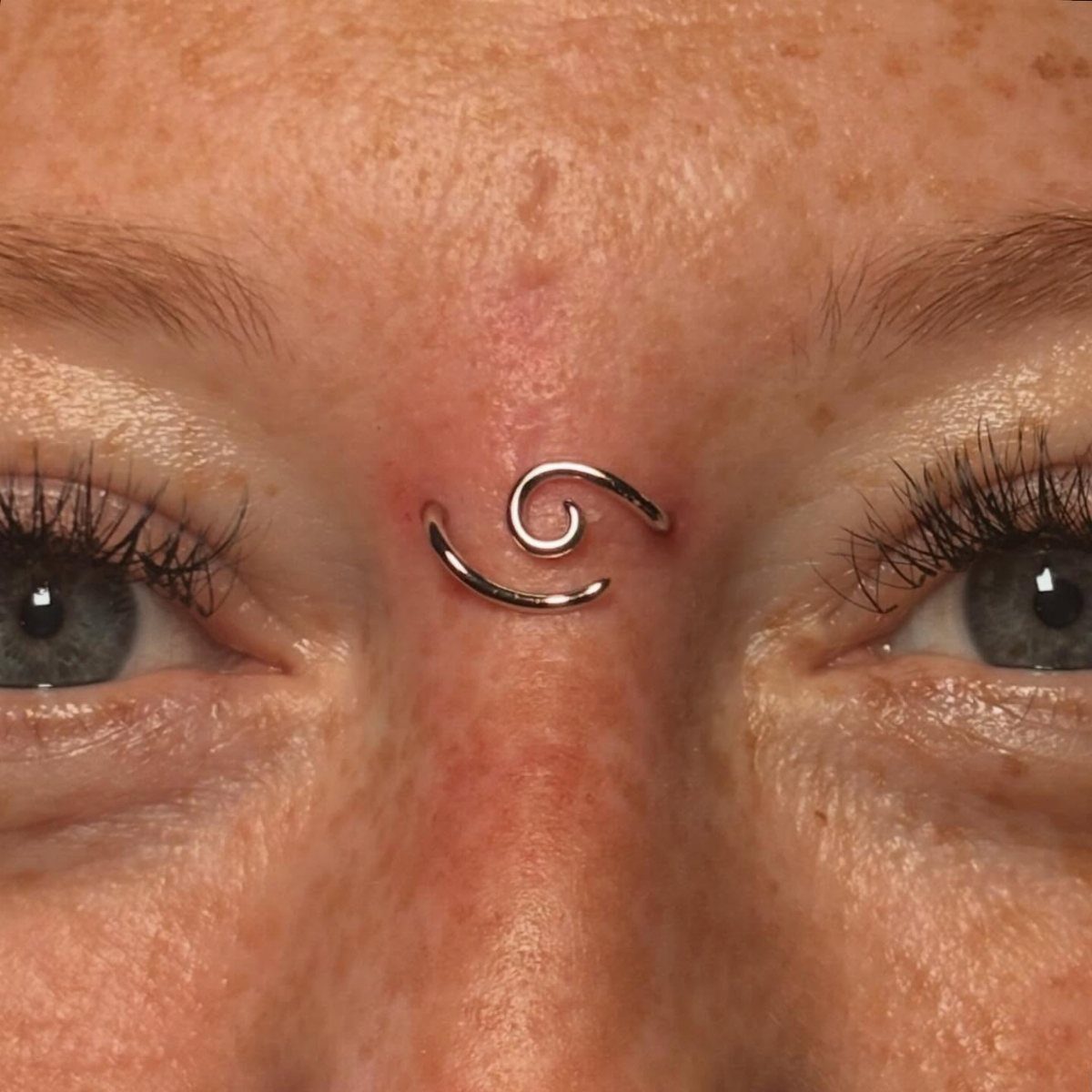 Piercing Between Eyebrows: 8 Styles and Placements 8 Silver spiral piercing centered between the eyebrows on a person's freckled skin.