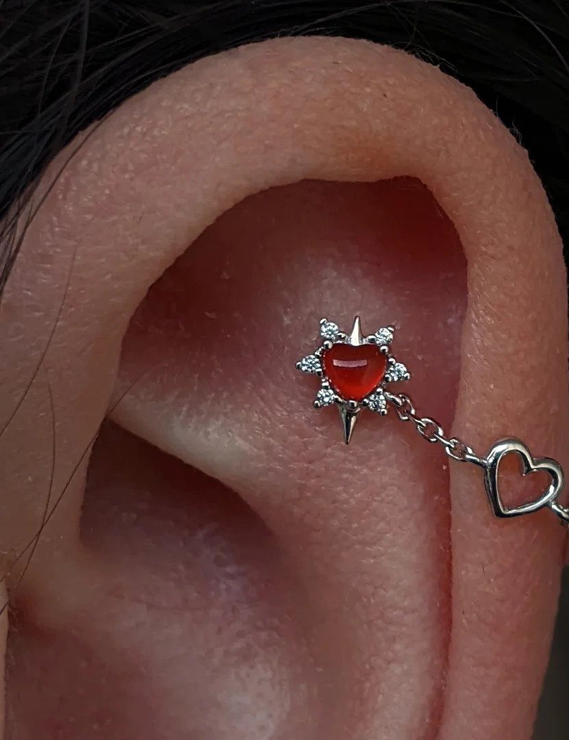 Human ear with a red heart star piercing and a connecting silver chain.
