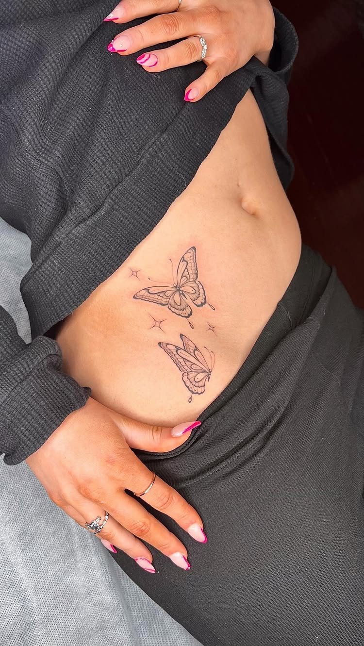 Two butterfly tattoos on a woman's hip and lower abdomen with pink nails.