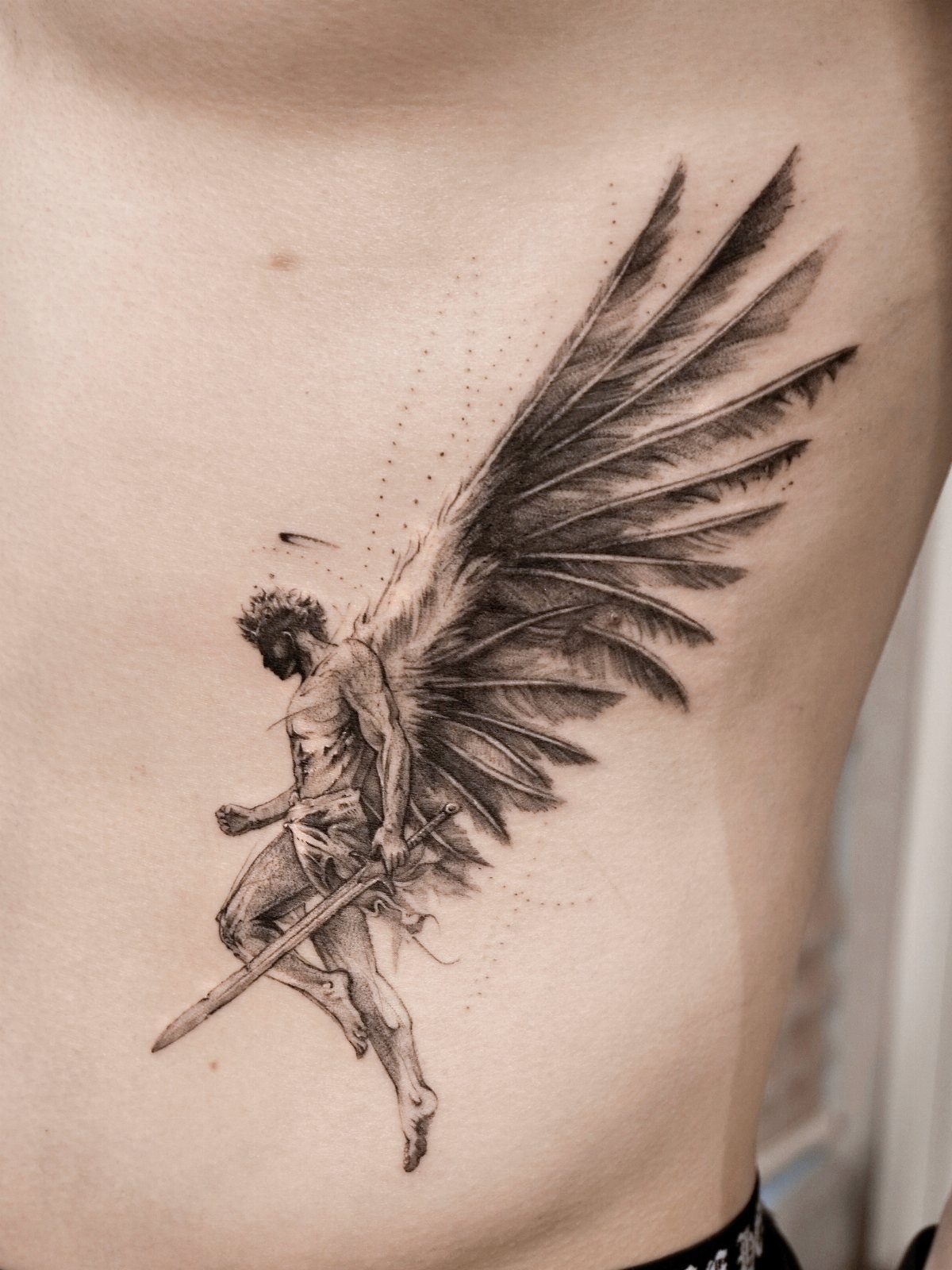 Winged figure tattoo with a sword on a person's ribcage in black and grey.