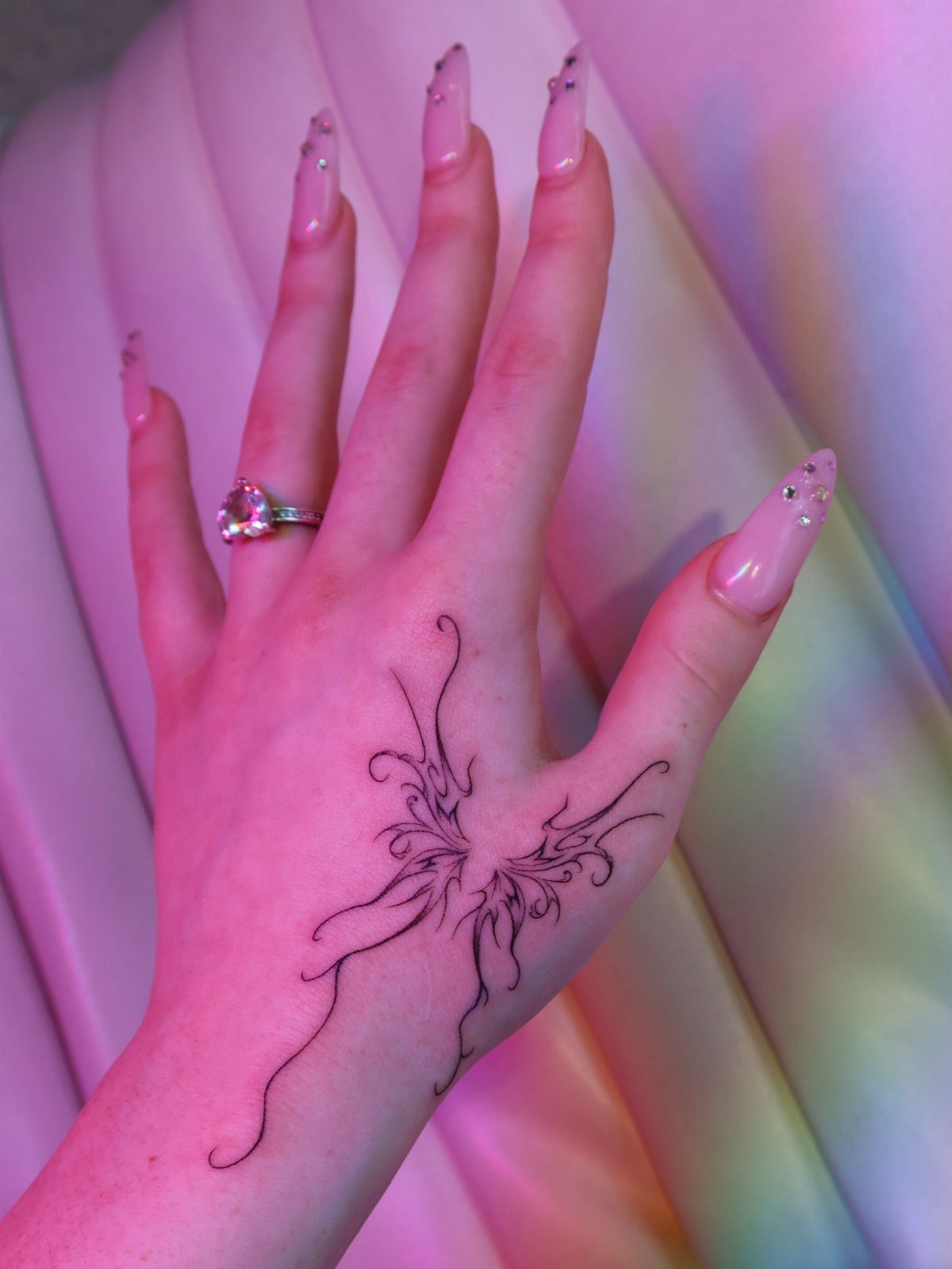 Hand with long pink nails and a black abstract butterfly tattoo on the back.