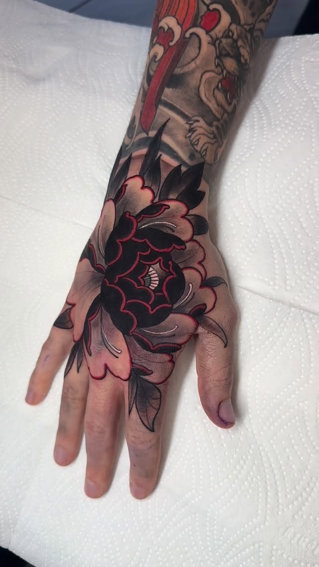 Large peony tattoo with black and red ink on the back of a hand.