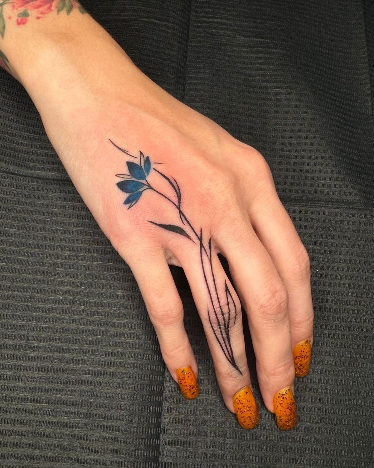 Hand with a blue flower tattoo and long orange speckled fingernails on dark background