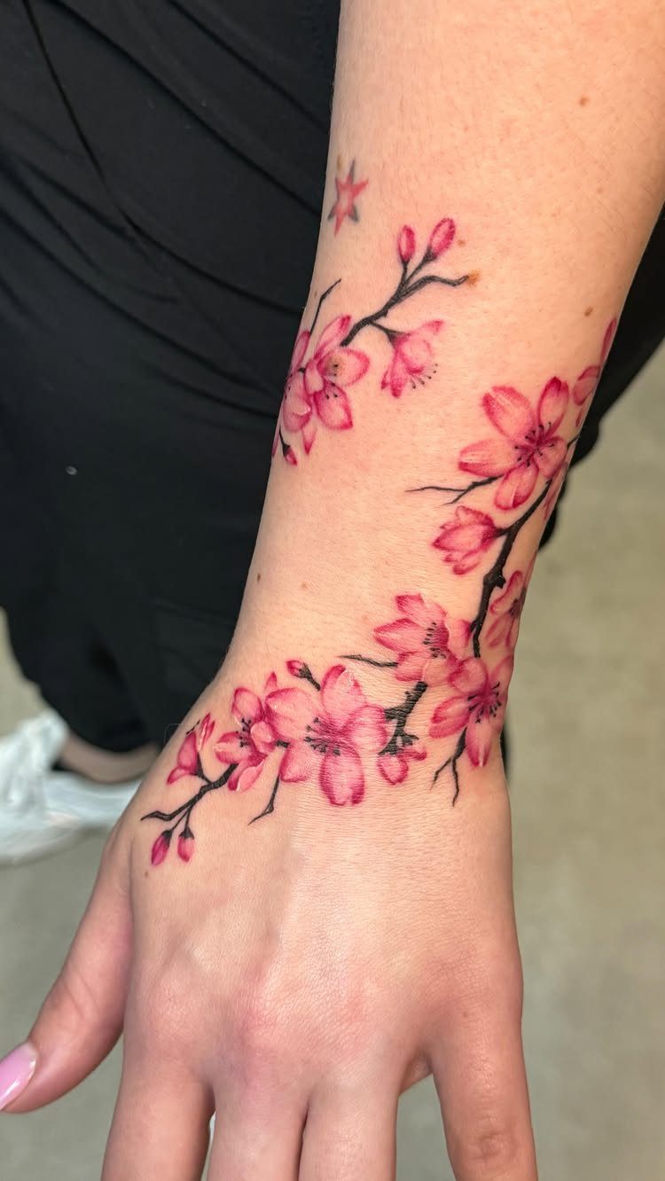 Cherry blossom branch tattoo with pink petals sweeping across a person's hand and wrist.