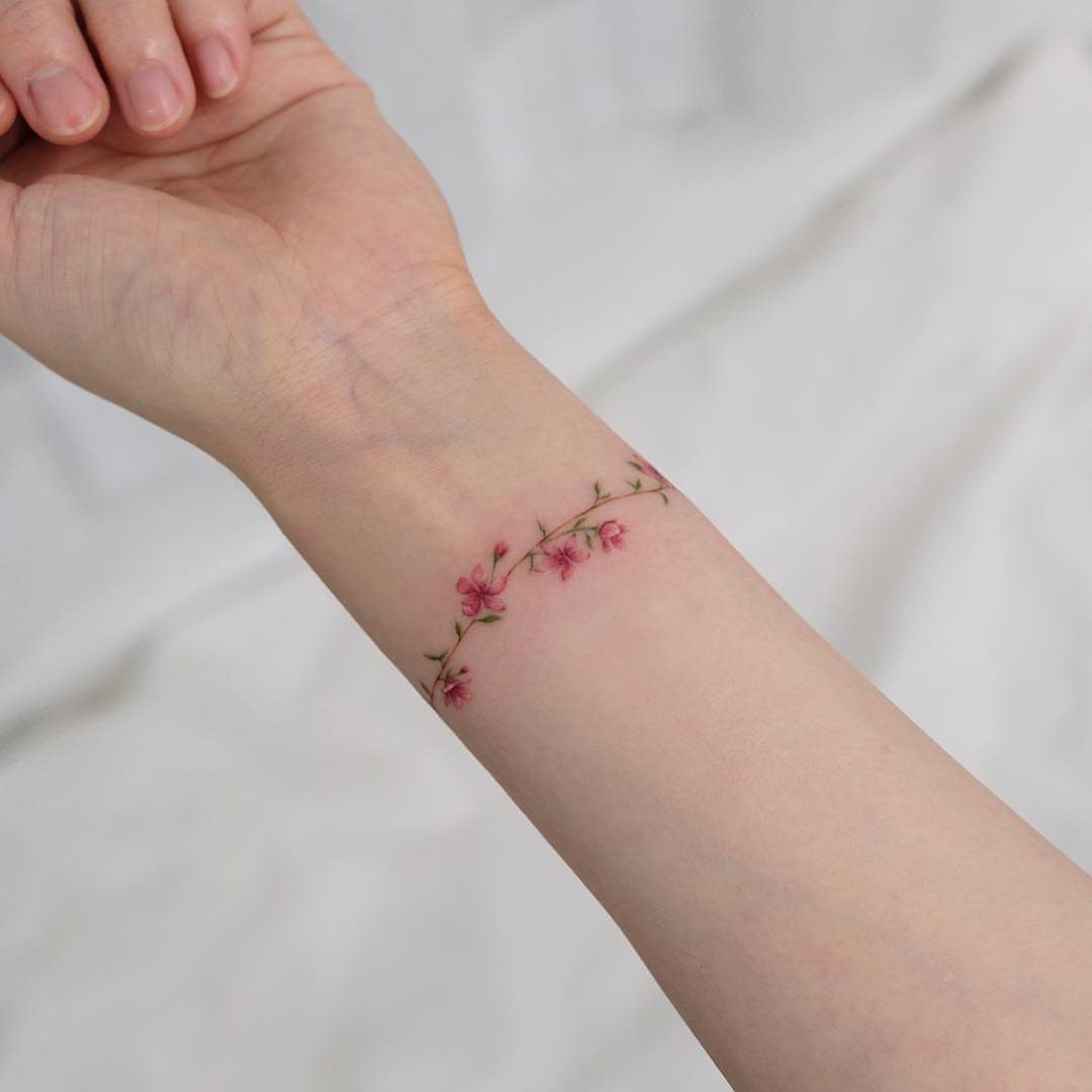 Wrist tattoo of a delicate green vine with small pink blossoms on skin.