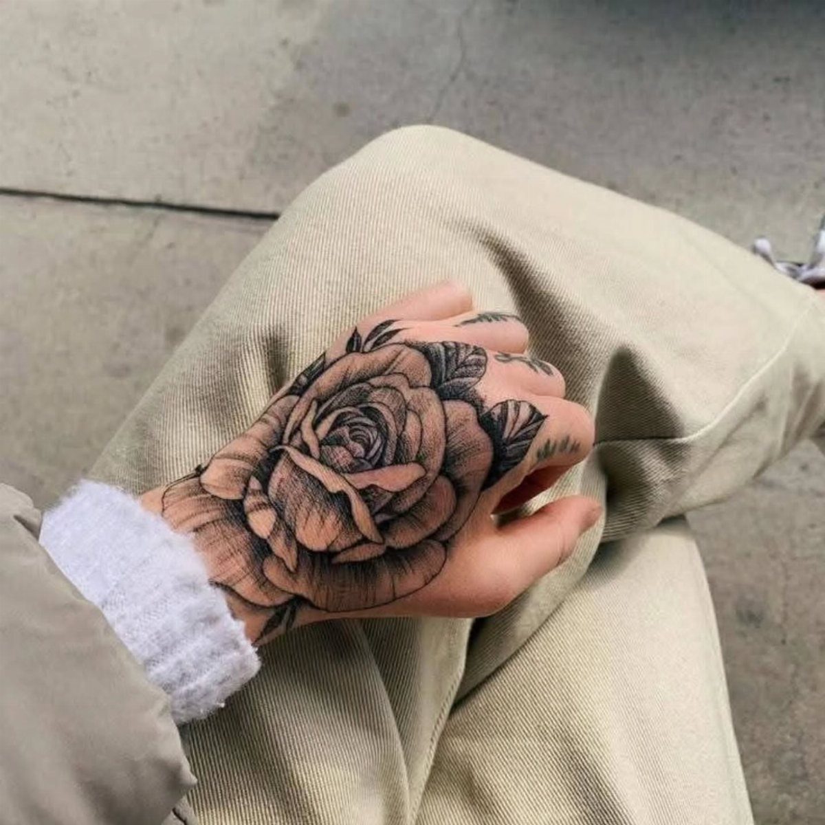 Hand with a detailed black and grey rose tattoo resting on beige trousers.