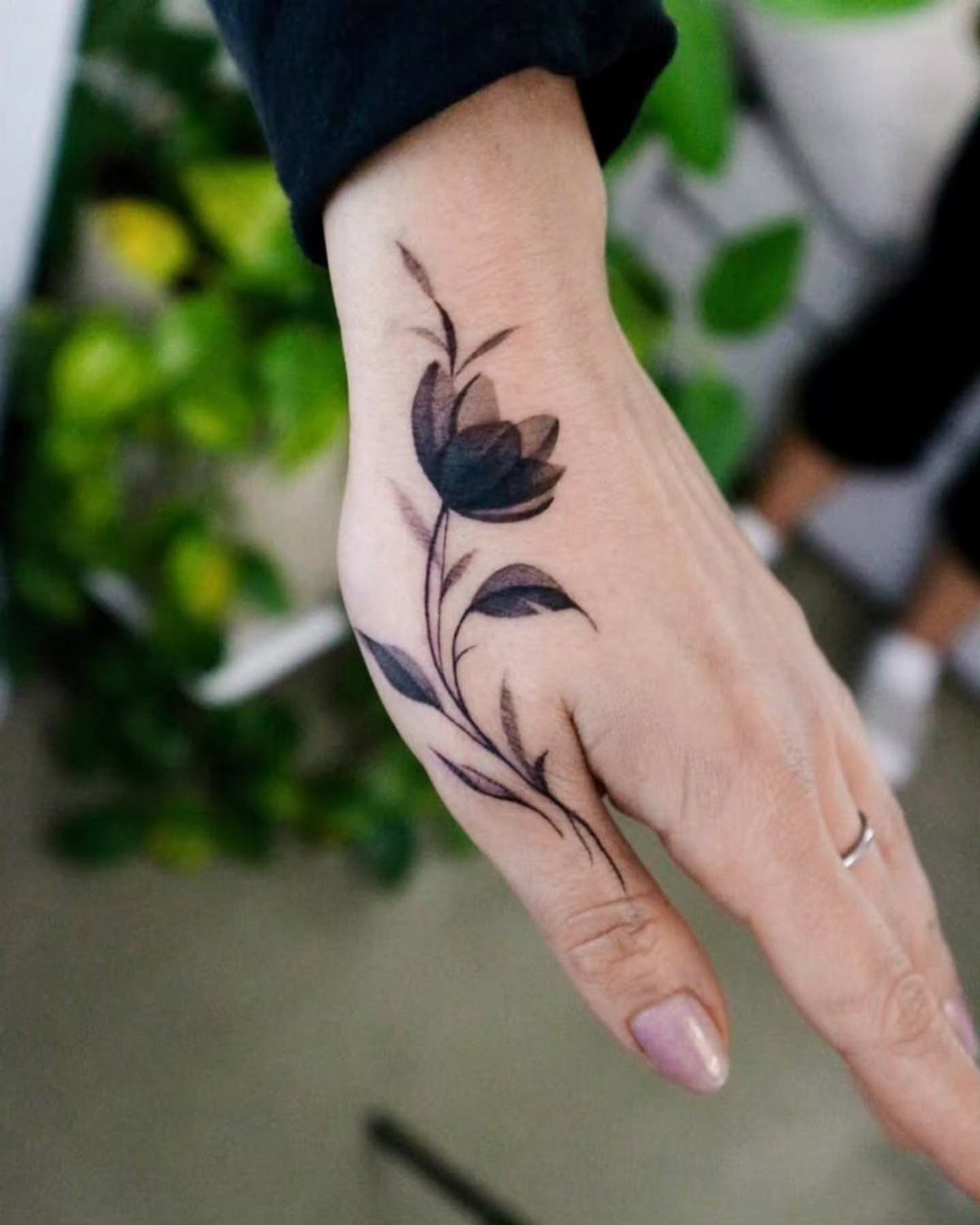 Black ink flower tattoo with a stem and leaves on a hand.