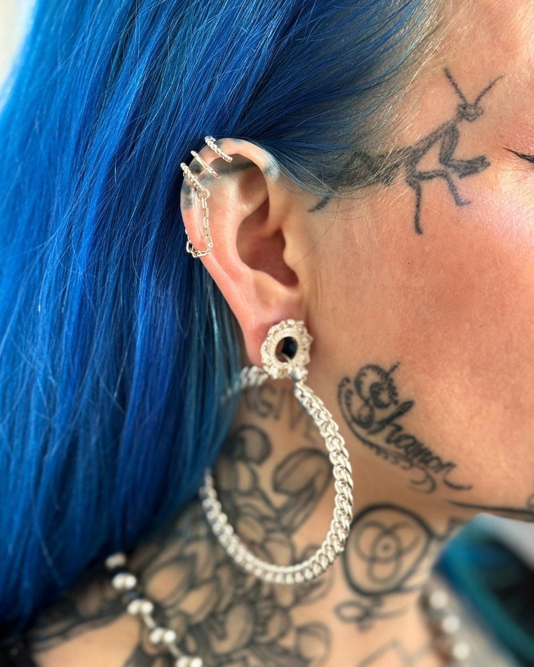 Ear with blue hair and tattoos featuring a chain piercing and large hoop earring.