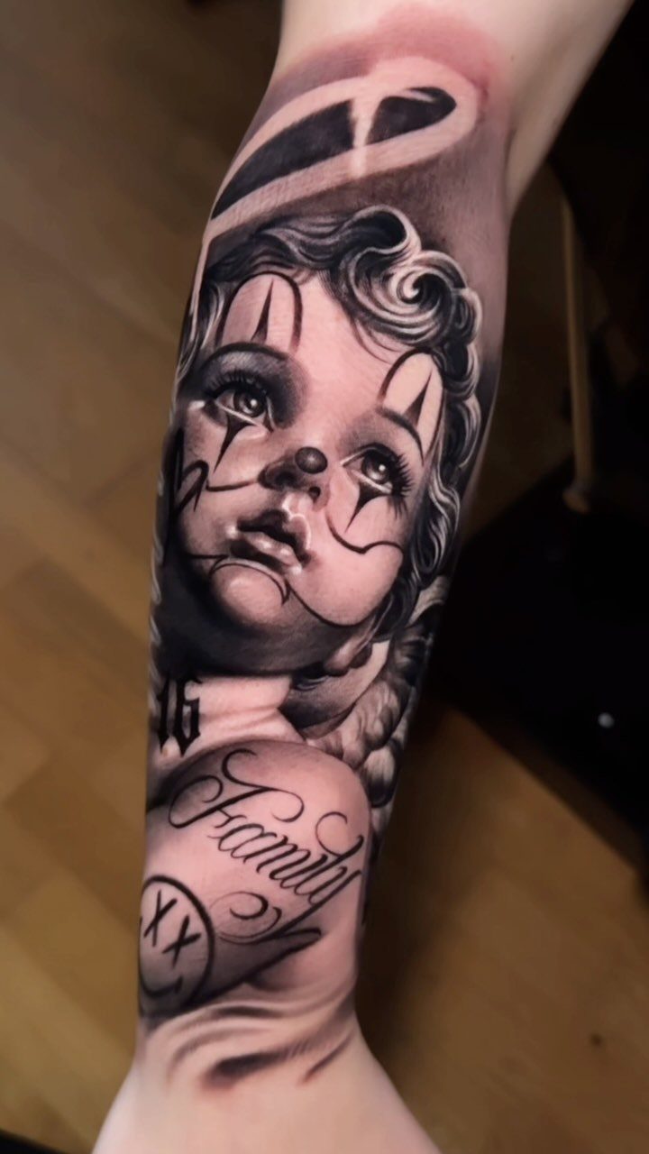 Cherub tattoo with clown makeup and the word Family on a forearm.