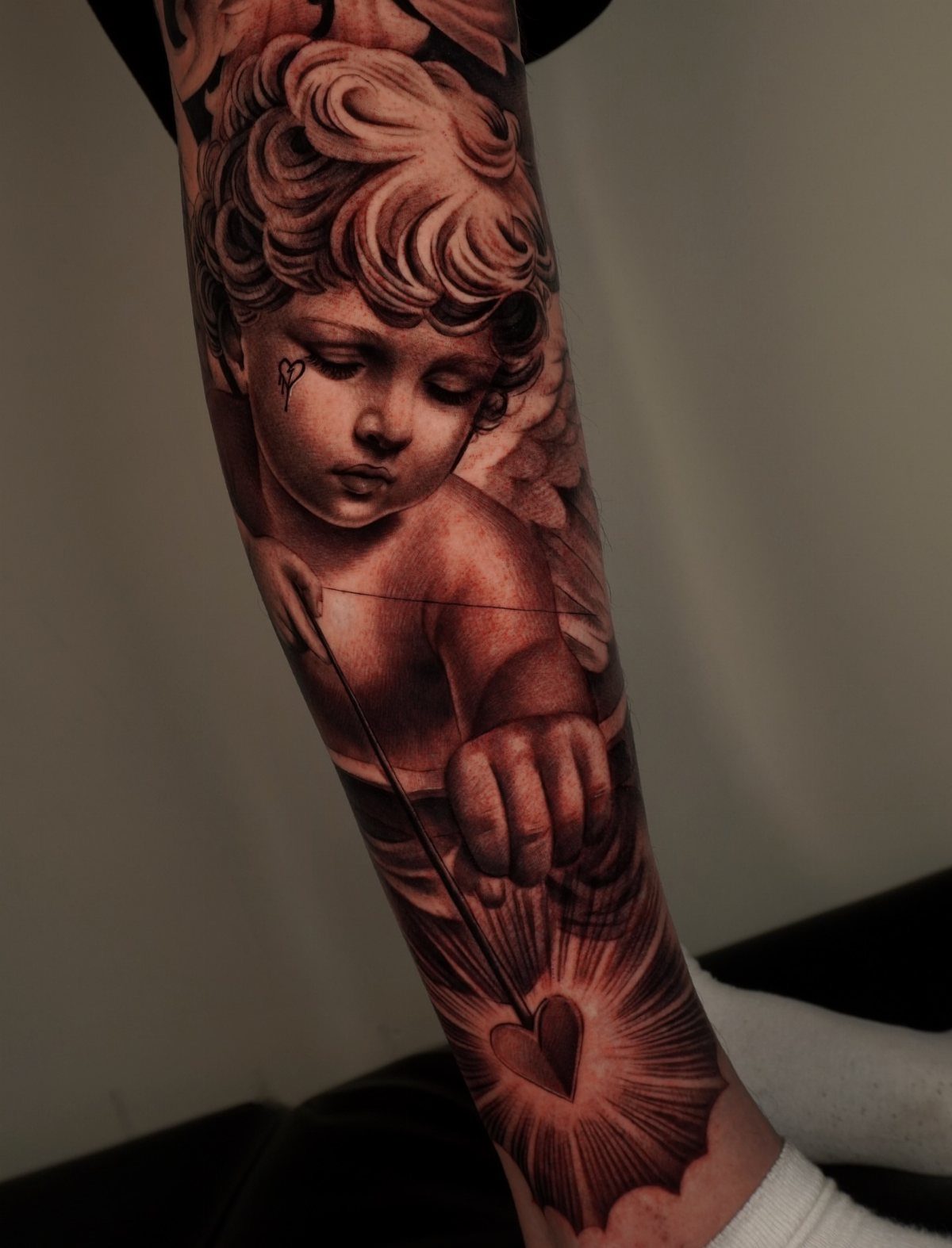 Cherub Cupid tattoo on an arm featuring a bow and arrow hitting a heart.