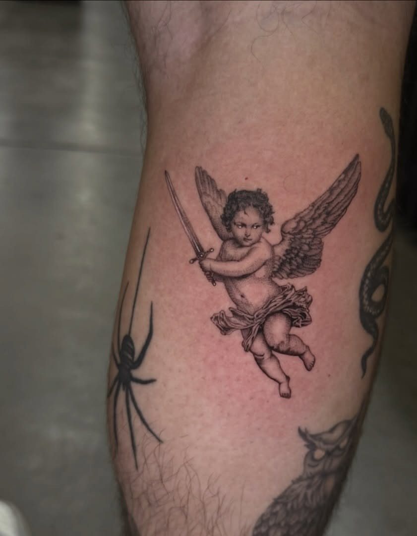 Realistic cherub tattoo holding a sword on a person's leg with other tattoos.