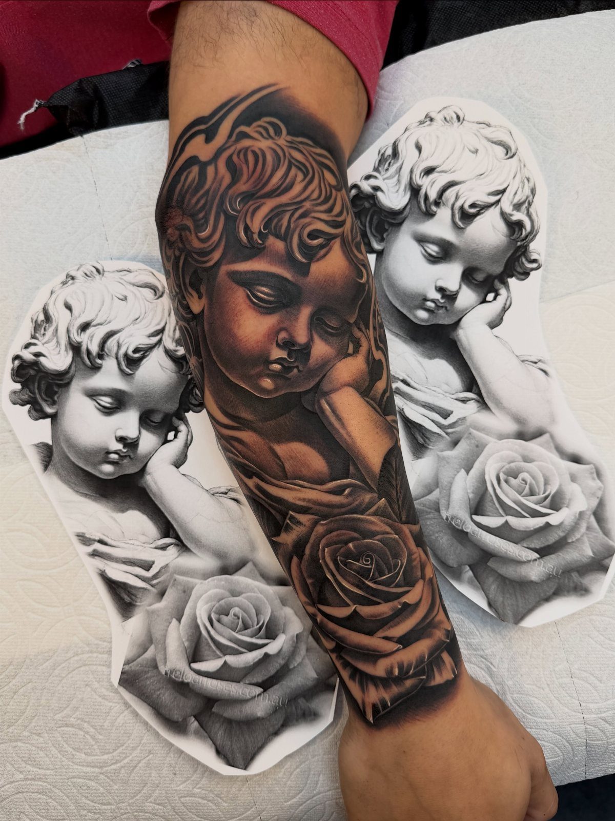 Realistic cherub tattoo on a forearm with accompanying printed reference sketches and roses.