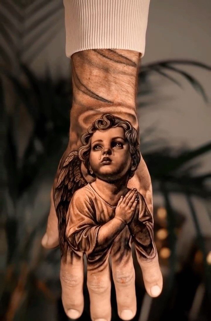 Cherub tattoo with praying hands and wings on a person's hand and wrist.