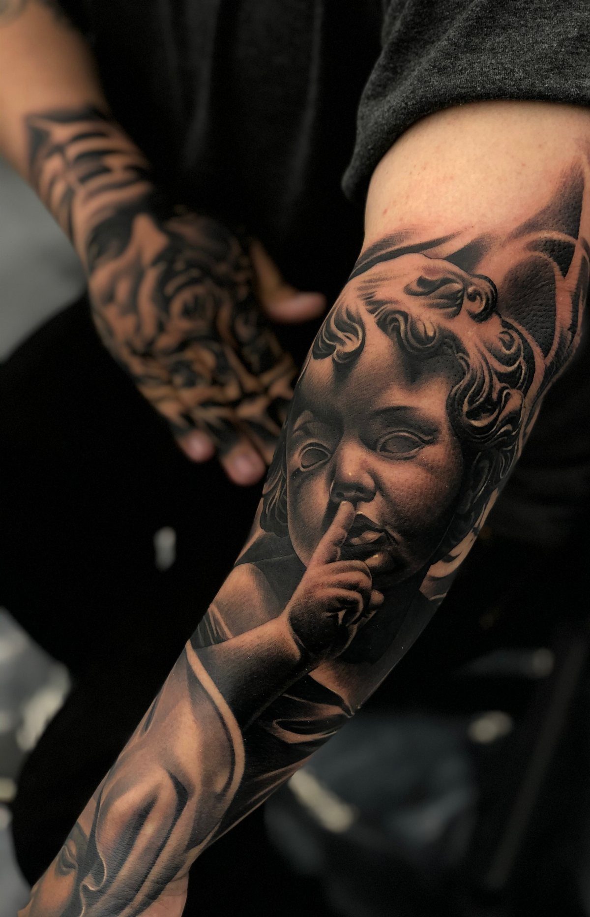 Realistic black and grey cherub tattoo on a person's forearm with a shushing gesture.
