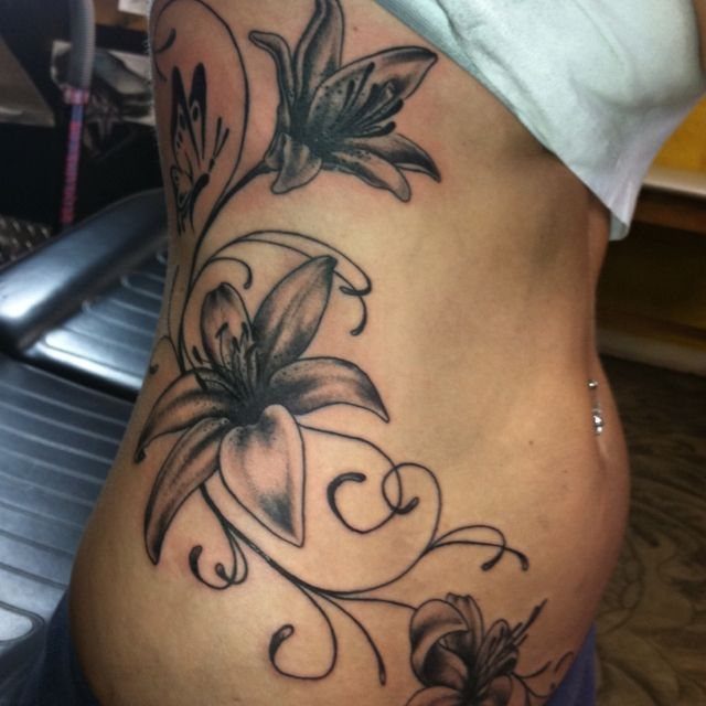 Rib Tattoos for Women: 9 Floral, Celestial and Script Ideas 3 Ribcage tattoo featuring lilies, swirling vines, and a small butterfly in black ink.
