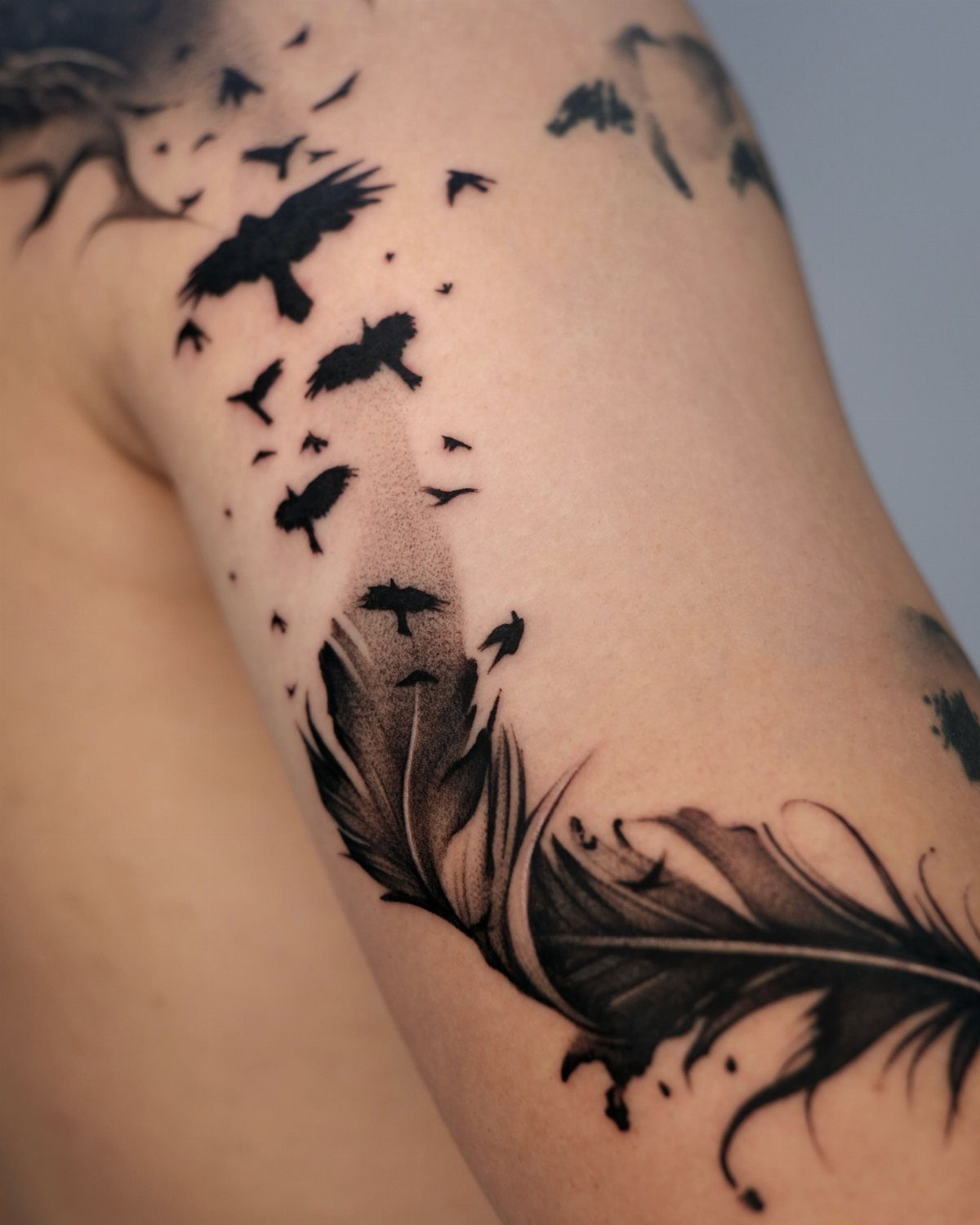 Black ink tattoo of a detailed feather with birds flying upwards on an arm.